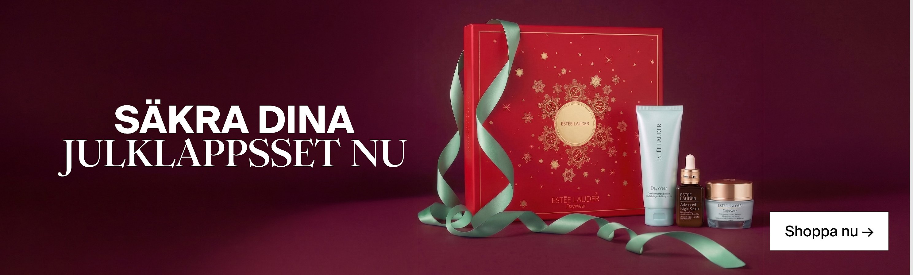 En röd presentask från Estée Lauder med guldfärgade snöflingor, ett grönt band och tre hudvårdsprodukter mot en mörkröd bakgrund.