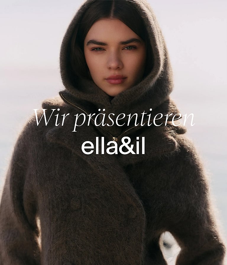 Eine lächelnde Frau in einem roten Strickoberteil isst eine Beere und hält eine gemusterte Schüssel in einer Küche. Text: Weiche Strickwaren für den Alltag.